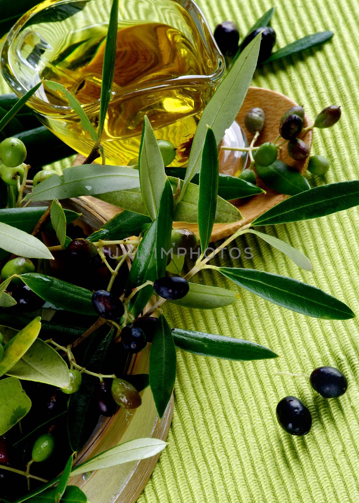 Olive Oil in Glass Gravy into Branches of Raw Green and Black Olives with Leafs in Wooden Bowl closeup on Green Textile background