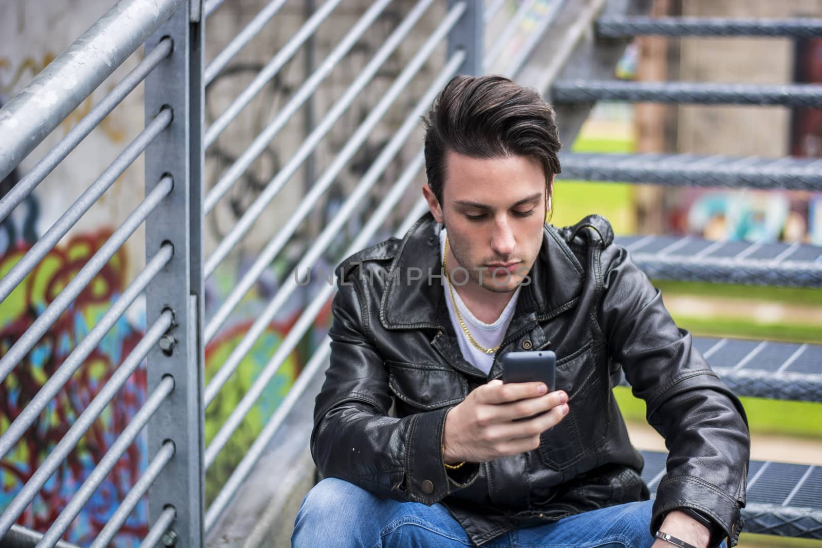 Handsome young man standing outdoors in urban environment on metal stairs, using cell phone