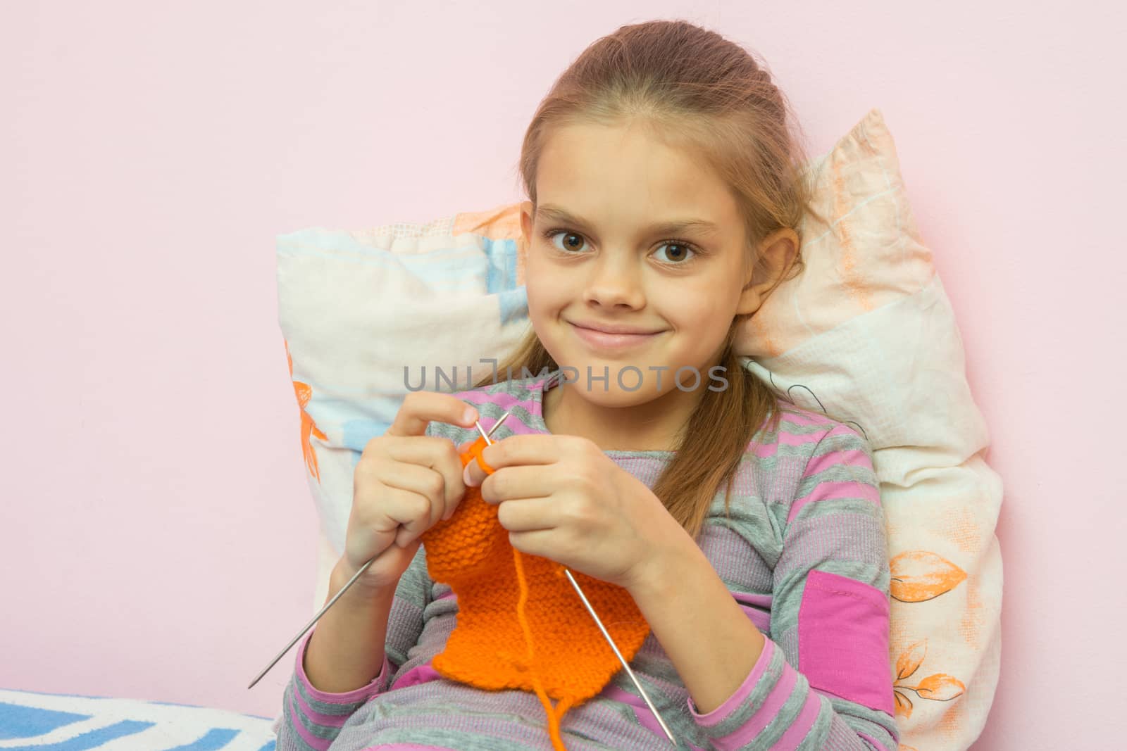 The girl knits on the needles orange scarf, smiling looked into the frame