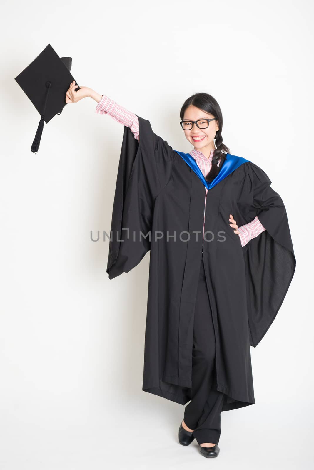 University student in graduation gown hand raised holding mortarboard. Full body portrait of east  Asian female model standing on plain background.