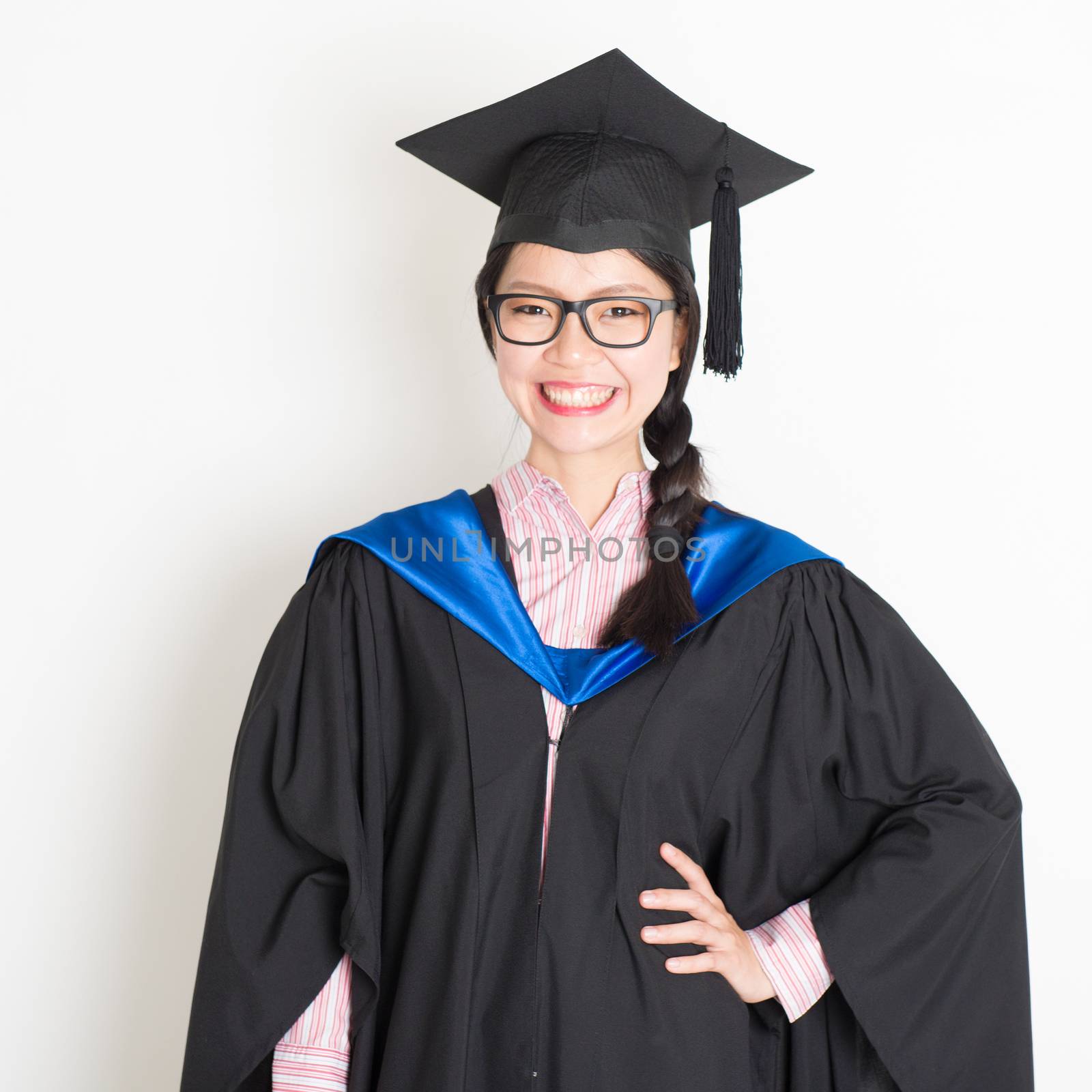 University student in graduation gown and cap. Portrait of east  Asian female model standing on plain background.