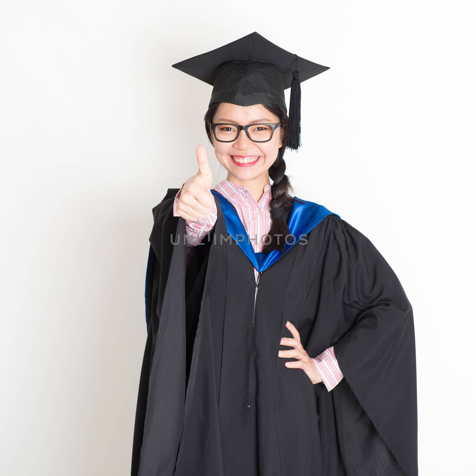 University student in graduation gown and cap giving thumb up sign. Portrait of east  Asian female model standing on plain background.