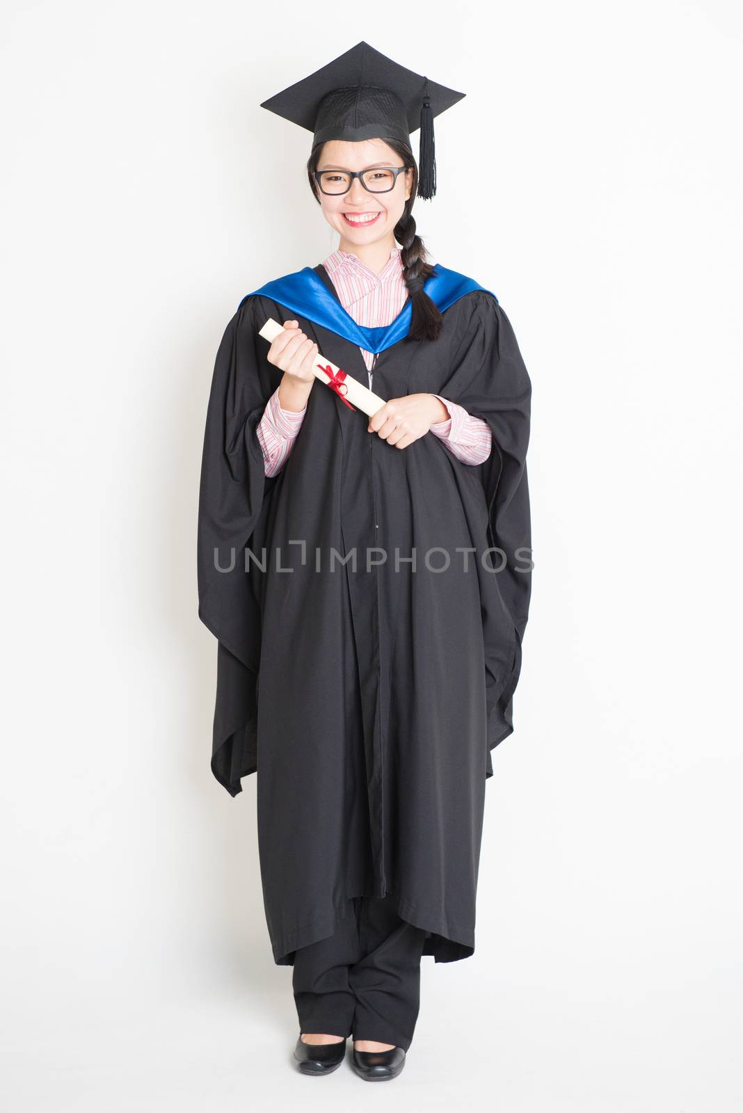 Happy university student in graduation gown and cap holding diploma certificate. Full body portrait of east  Asian female model standing on plain background.