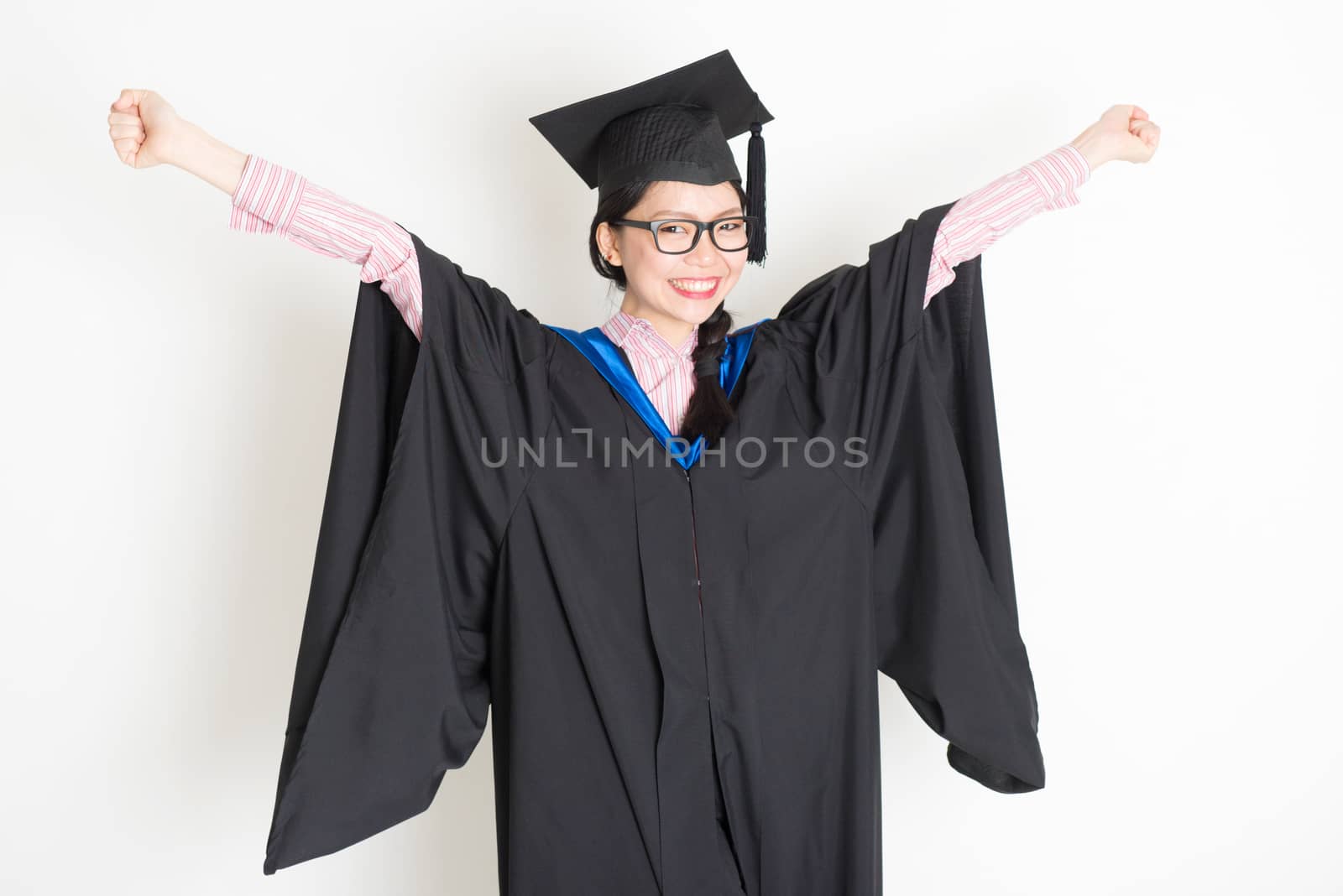 Happy university student in graduation gown and cap arms outstretched and smiling. Portrait of east  Asian female model standing on plain background.