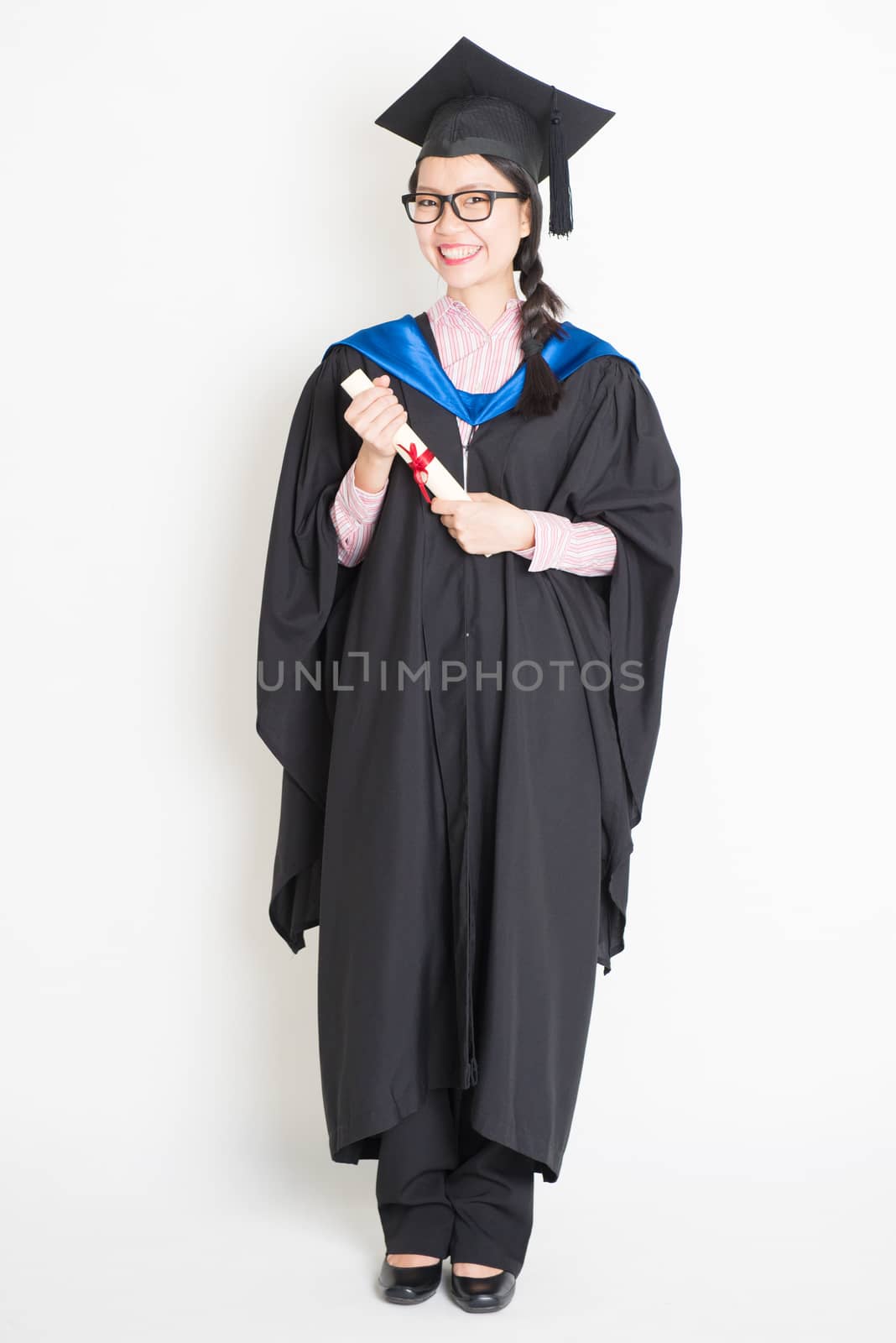 University student in graduation gown and cap holding diploma certificate. Full body portrait of east  Asian female model standing on plain background.