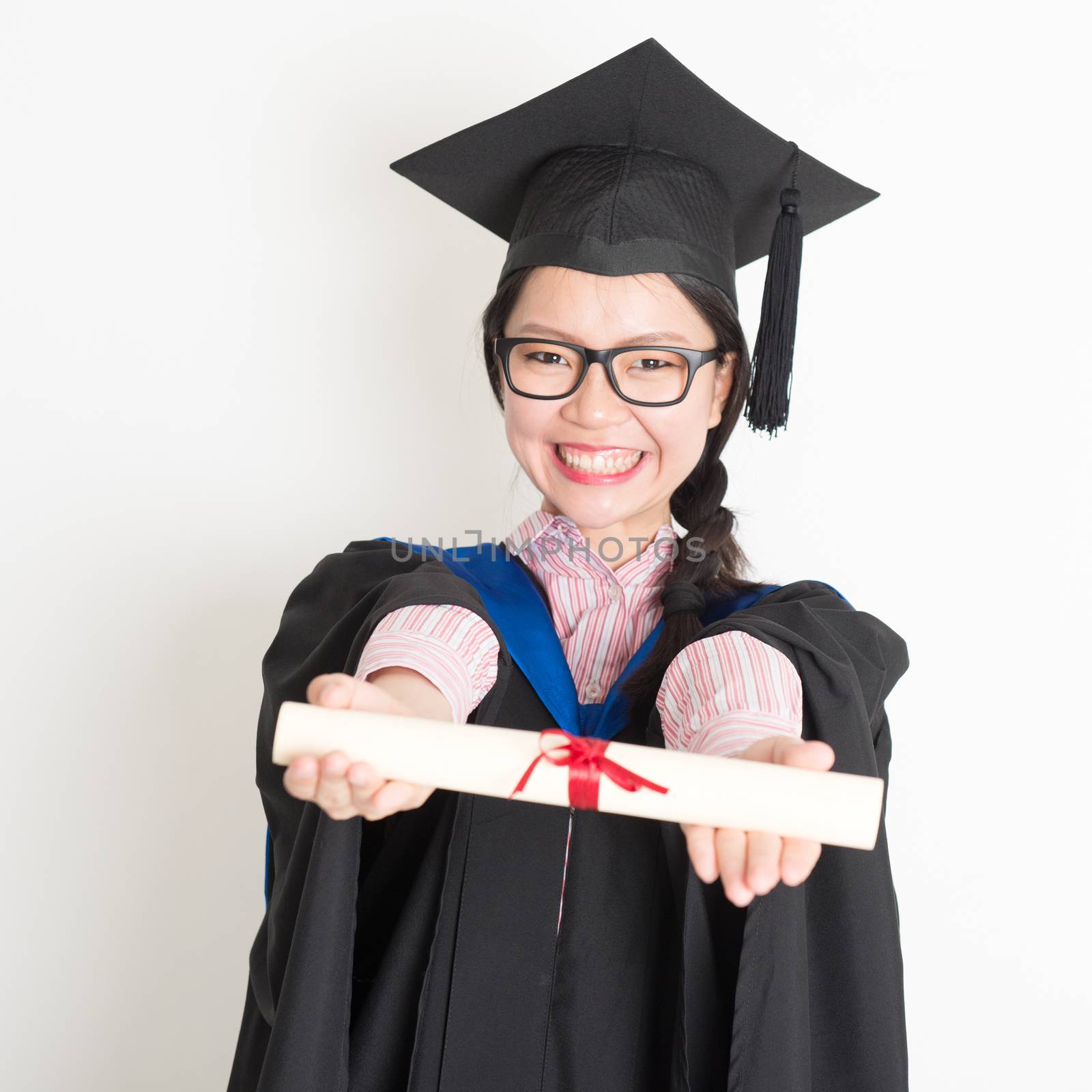 Happy university student in graduation gown and cap showing diploma certificate. Portrait of east  Asian female model standing on plain background.