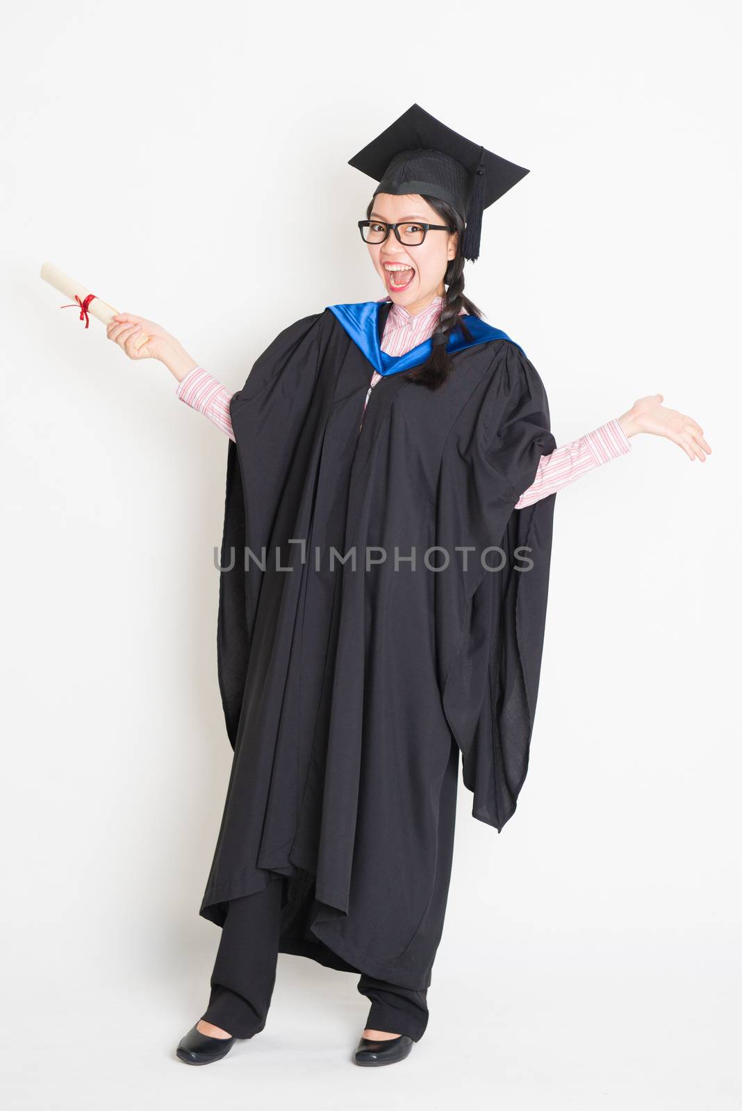 University student in graduation gown and cap holding diploma certificate laughing. Full body portrait of east  Asian female model standing on plain background.