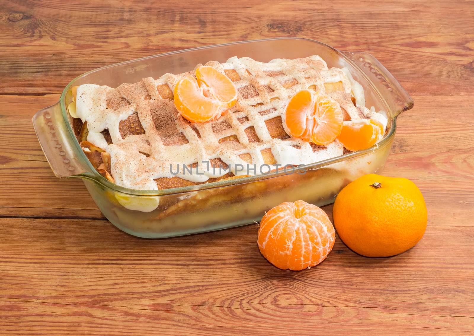 Sweet pancakes filled with cottage cheese with sour cream and sprinkled with cinnamon powder in the rectangular glass roasting pan and mandarins on old wooden surface
