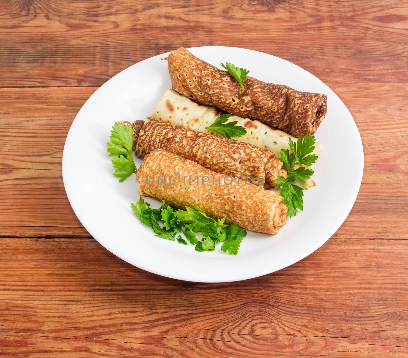 Pancakes filled with meat filling and parsley twigs on a white dish on an old wooden surface
