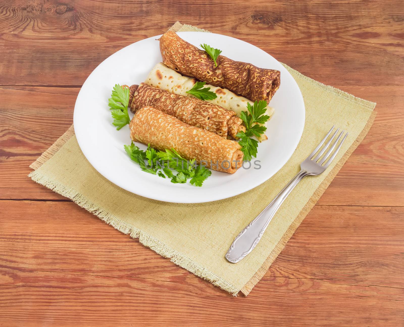 Pancakes filled with meat filling and parsley twigs on a white dish and fork on a cloth napkin on an old wooden surface
