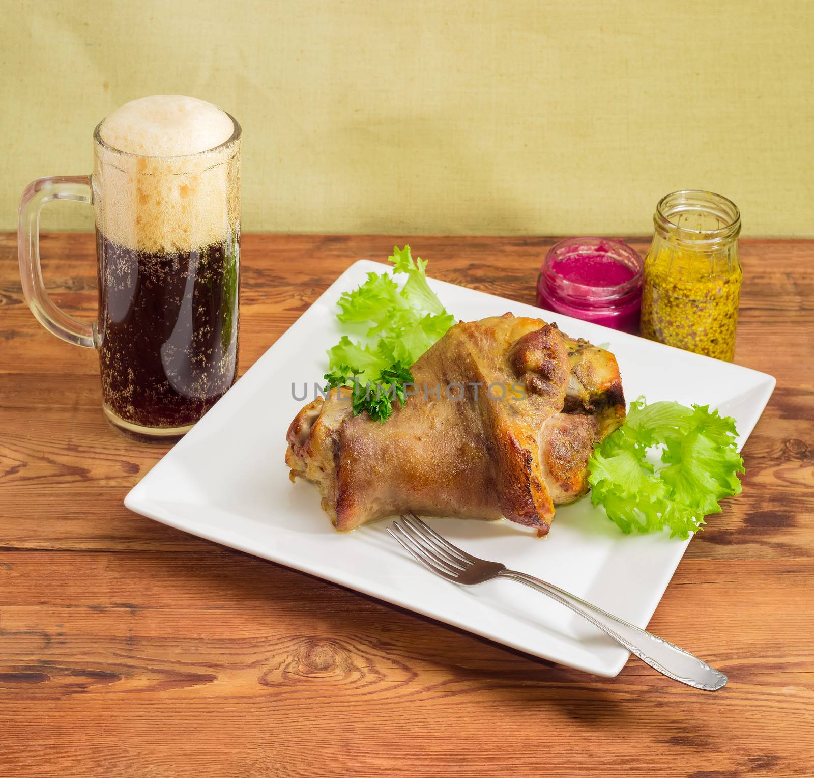 Baked ham hock with lettuce and parsley on a square white, glass of dark beer, condiment and fork on a surface of old wooden planks
