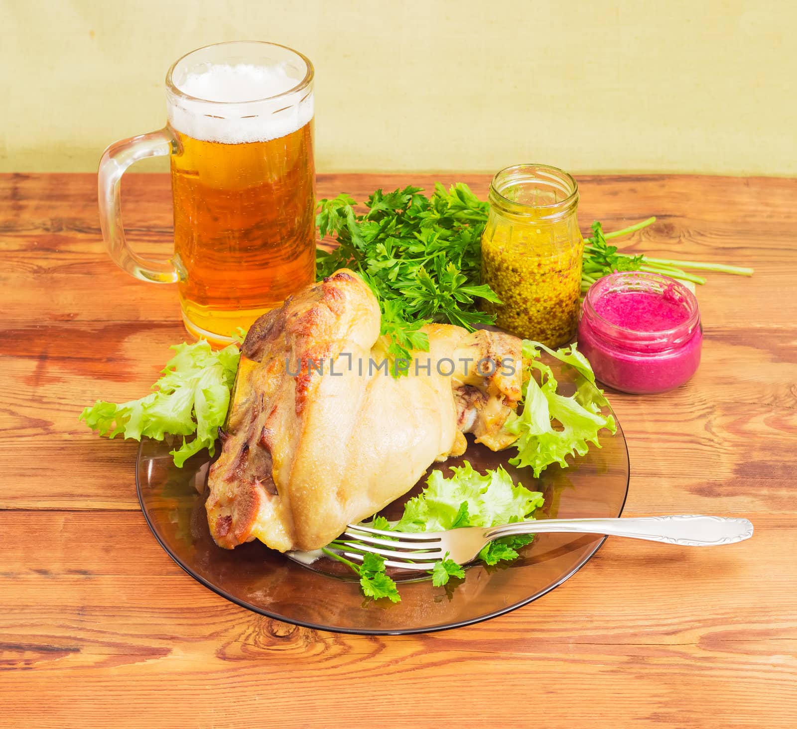 Baked ham hock with lettuce and parsley on a glass dish, glass of lager beer, beet horseradish sauce, French mustard, fork on an old wooden surface
