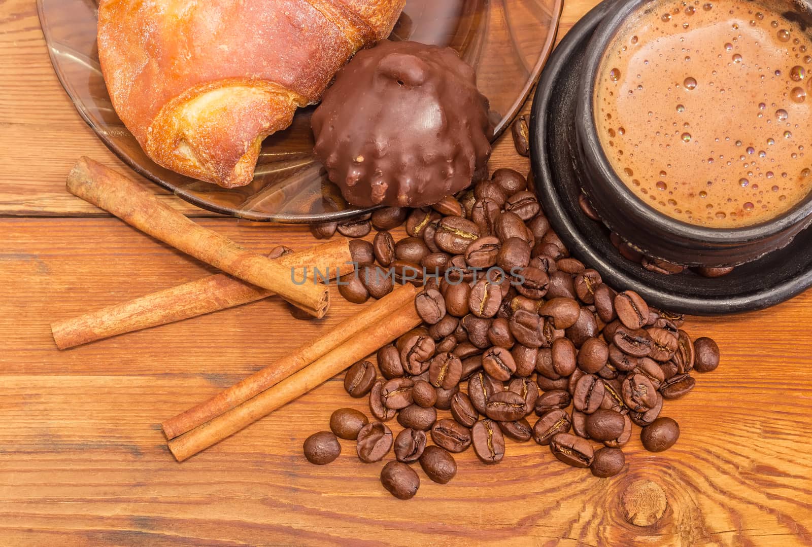 Fragment of black ceramic cup with freshly brewed coffee closeup on the background of roasted coffee beans, cinnamon sticks, chocolate truffle and croissant on a wooden surface
