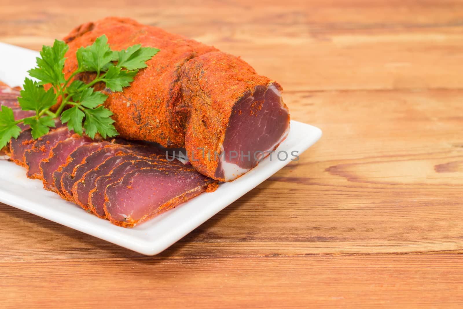 One piece and sliced dried pork tenderloin with twigs of parsley closeup on a white dish on a wooden surface
