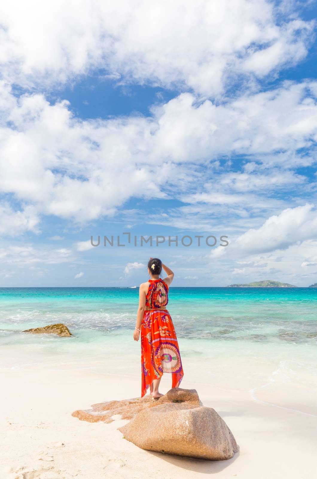 Traditinaly dressed local woman wearing long floral summer dress relaxing, doing yoga on Anse Patates beach, La Digue Island, Seychelles. Summer vacations on picture perfect tropical island concept.