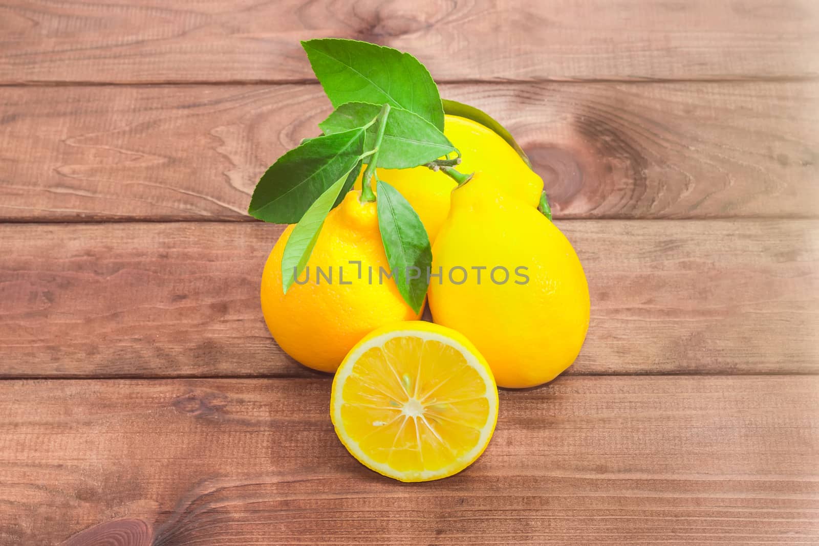 Several whole fresh yellow lemons with twigs and leaves and one lemon, cut in half on a dark wooden surface
