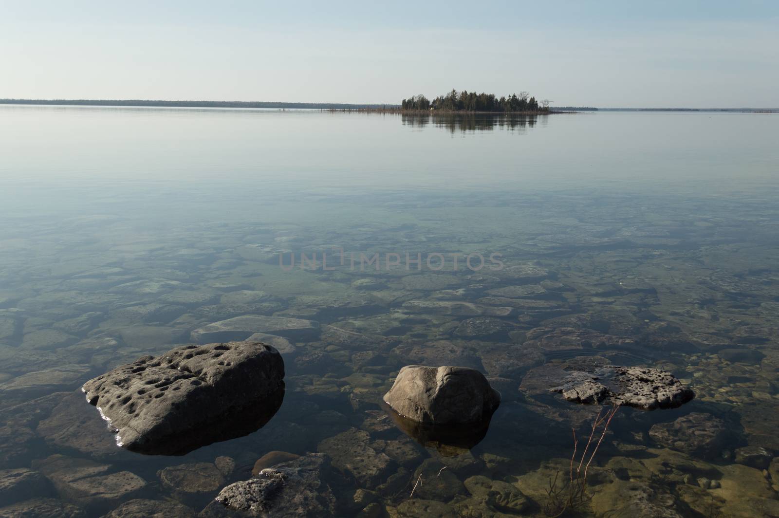 Dead calm afternoon image of shallow and clear lake huron water and limestone rocks along theshoreline.  A small tree-covered island is on the horizon. Overall feeling is a calm, peaceful, tranquil, serene and wild background.