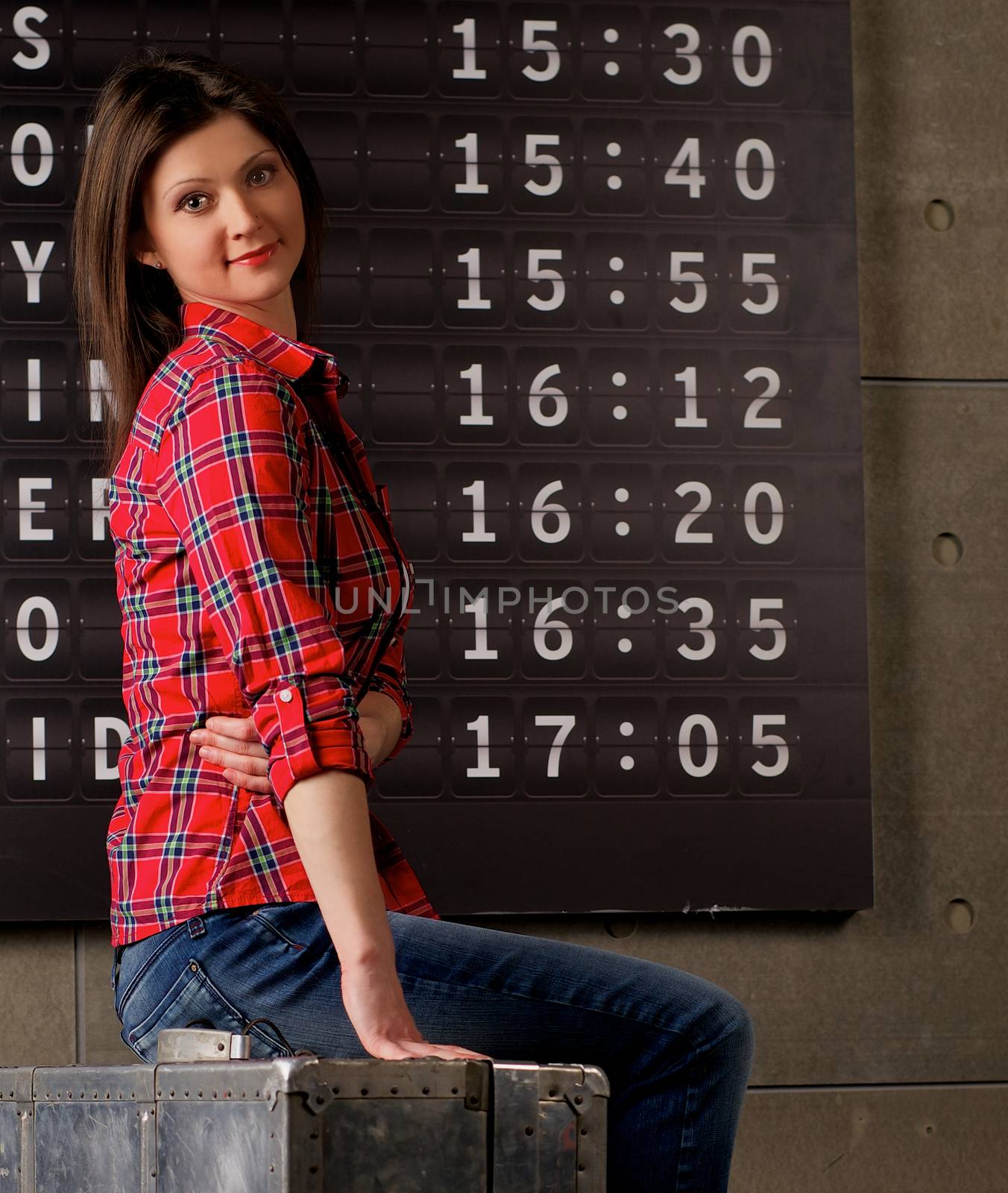 Attractive Young Woman in Checkered Shirt and Jeans Sitting on Obsolete Suitcase against Arrival Departure Board