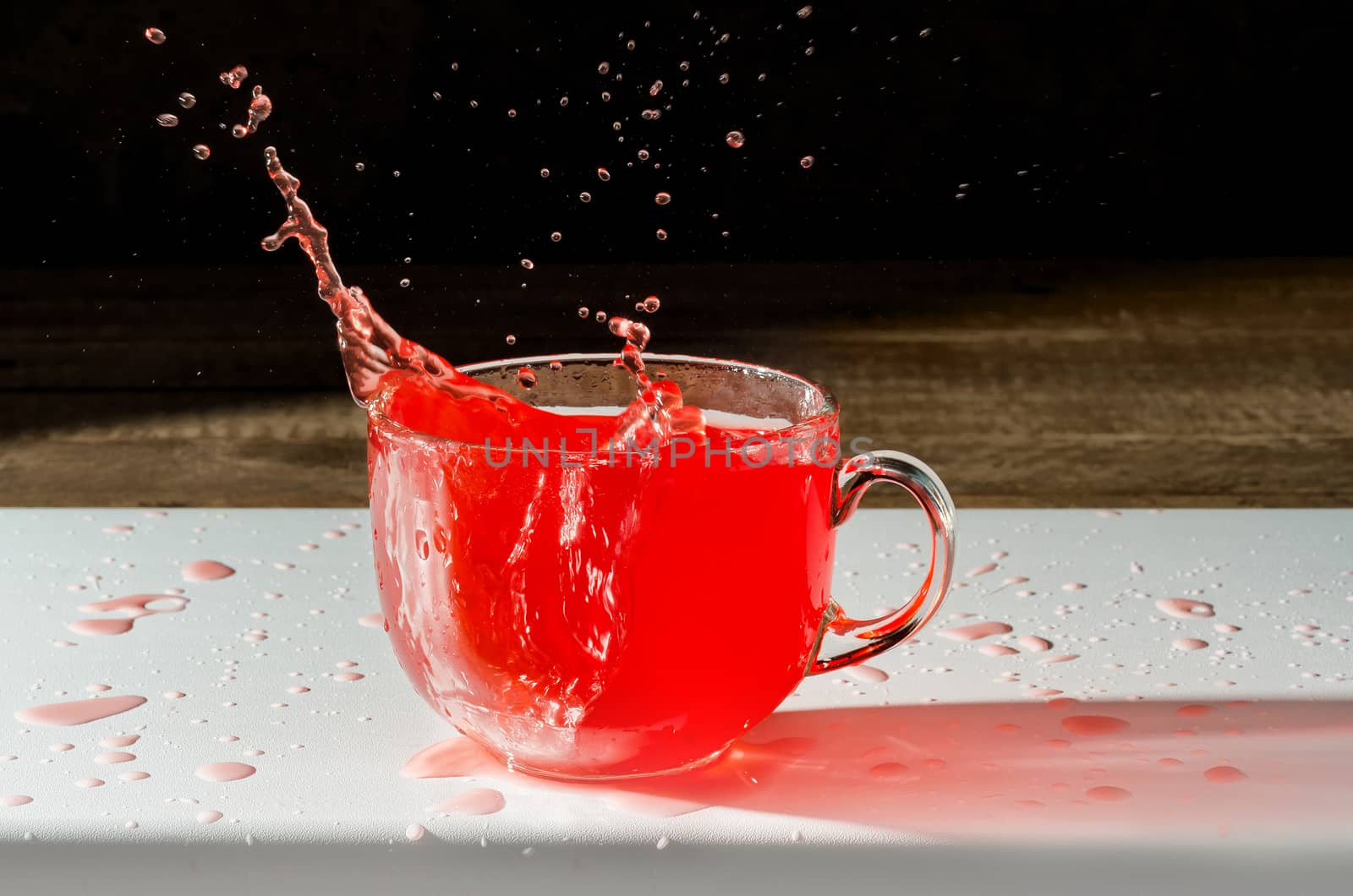 Juice in a Cup and splash with spray on the dark background. White surface, spilled drops and reflection.