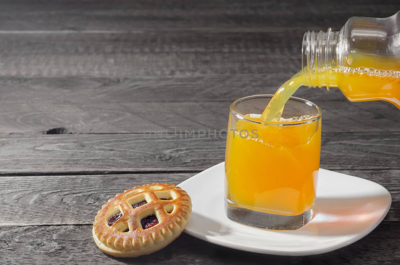 Cookies and orange juice poured from a bottle into a glass on gray wooden background. Selective focus.