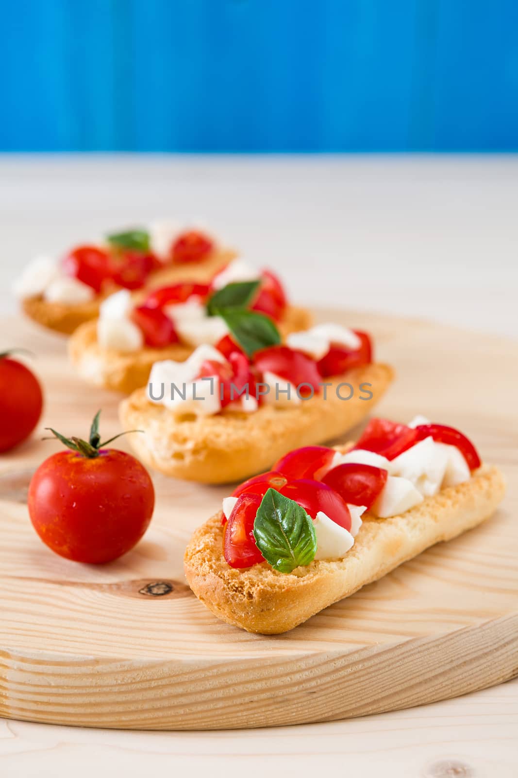 Italian crostini with mozzarella, cherry tomatoes and basil over a chopping board and blue background