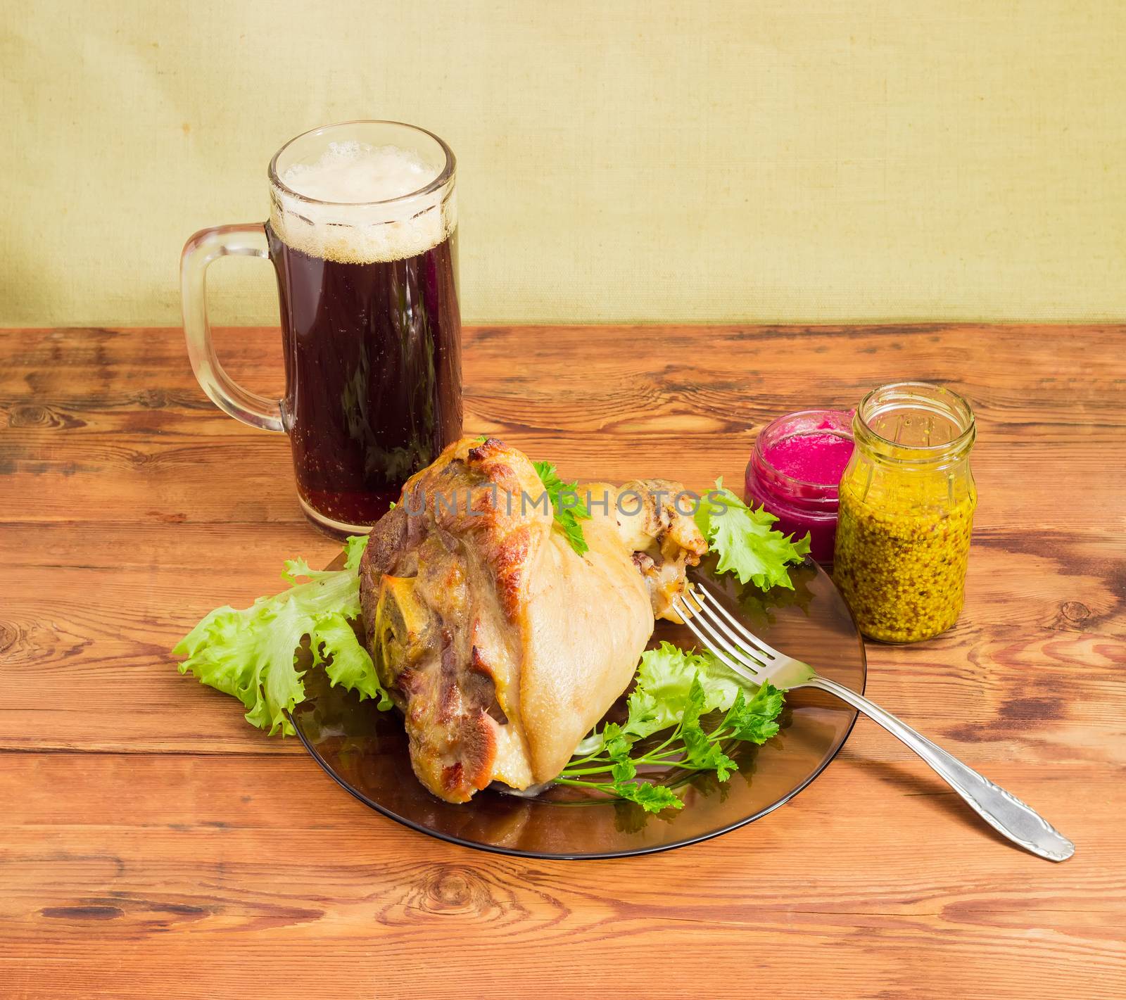 Baked ham hock with lettuce and parsley on a glass dish, glass of dark beer, beet horseradish sauce, French mustard and fork on a surface of old wooden planks
