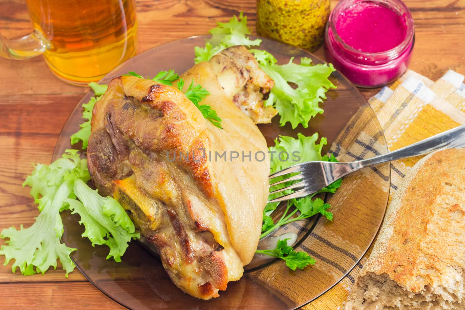 Baked ham hock with lettuce and parsley and fork on a glass dish on the background of a beer and condiments closeup
