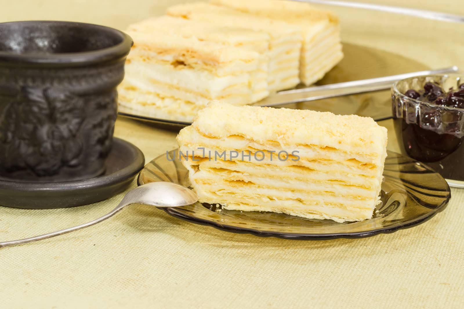 Piece of layered sponge cake on a glass saucer with spoon on the background of the rest of the cake, black cup and jam closeup on a cloth surface
