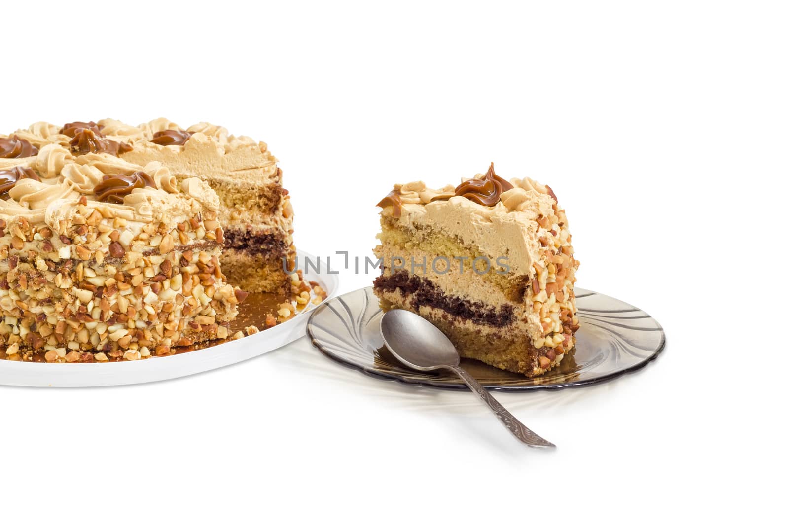 Piece of layered sponge cake, decorated with butter cream, caramelized condensed milk and nuts with spoon on a glass saucer and partly sliced cake on a light background
