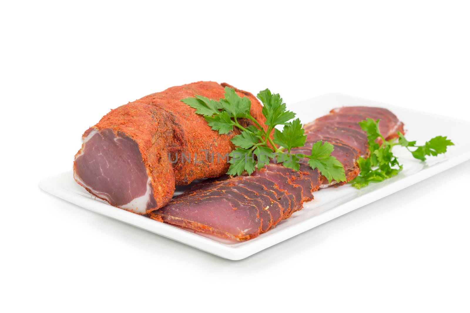 One piece and sliced dried pork tenderloin with twigs of parsley on a white rectangular dish closeup on a light background 
