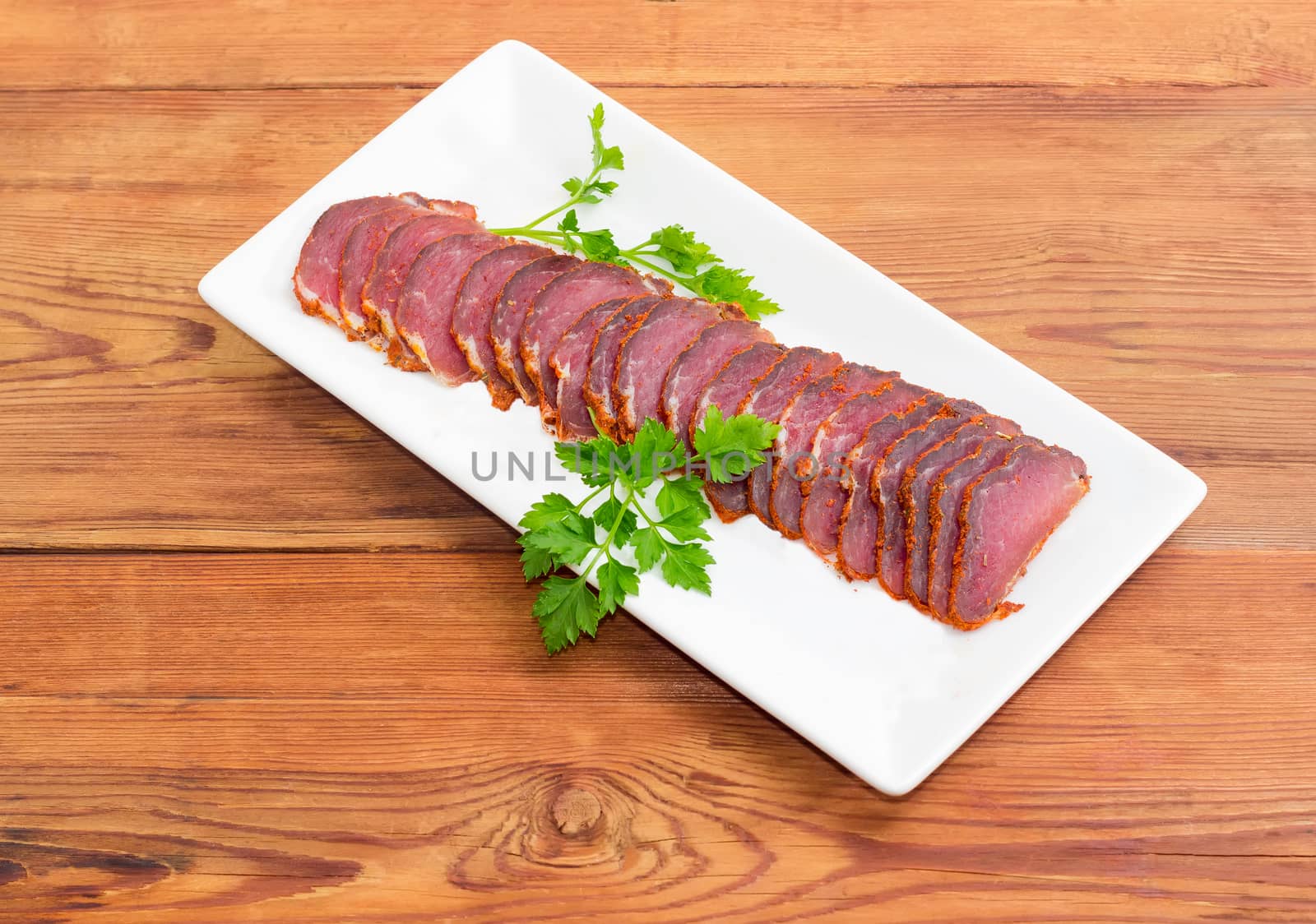 Sliced dried pork tenderloin with twigs of parsley on a white rectangular dish on a wooden surface
