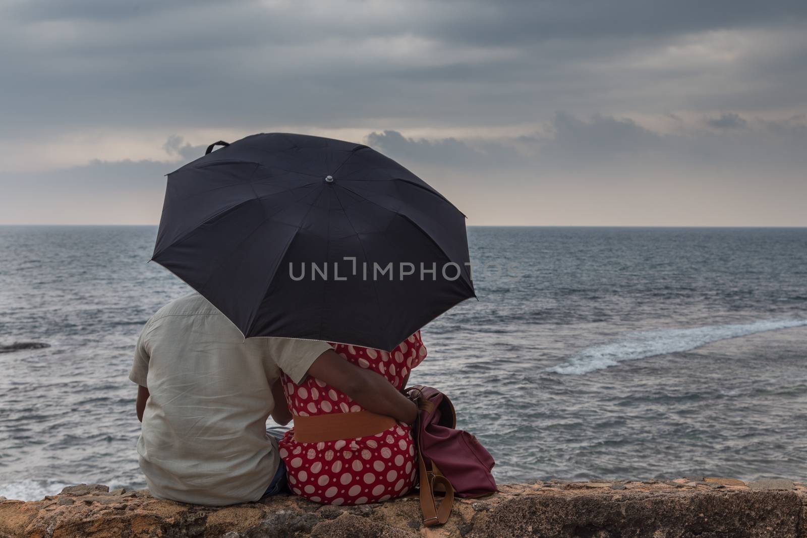 Rear View of a Indian couple sitting by sea with an umbrella looking at storm on ocean horizon.