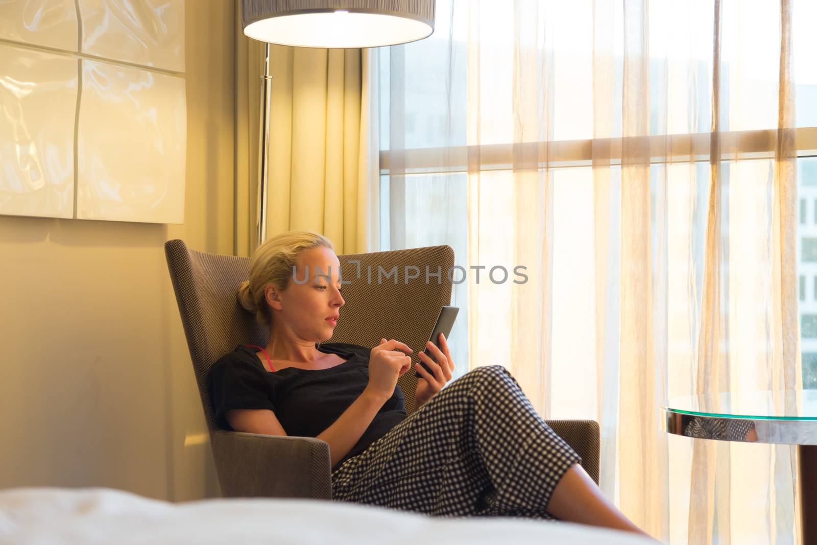 Casually dressed attractive blonde caucasian woman holding and using smart phone while sitting on armchair in modern hotel room.