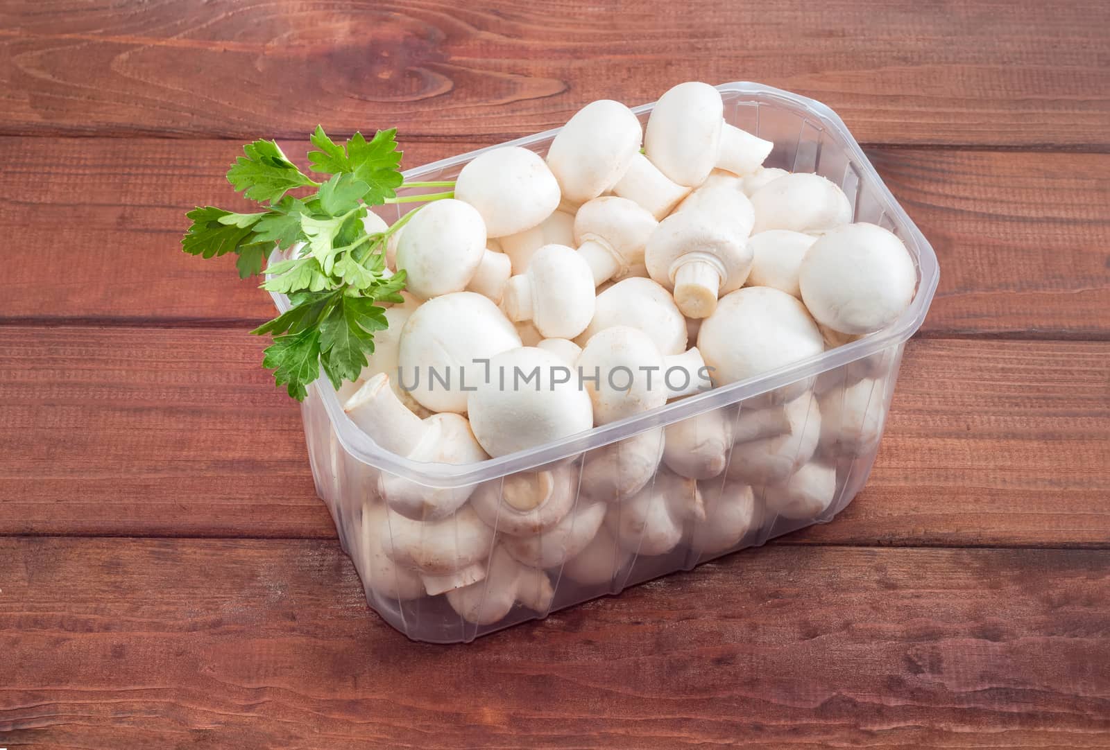 Fresh uncooked button mushrooms in a transparent plastic tray and sprig of parsley on a dark wooden surface
