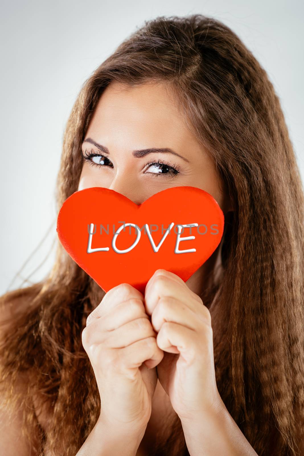 Beautiful smiling girl hiding behind a red heart that says Love. Looking at camera.