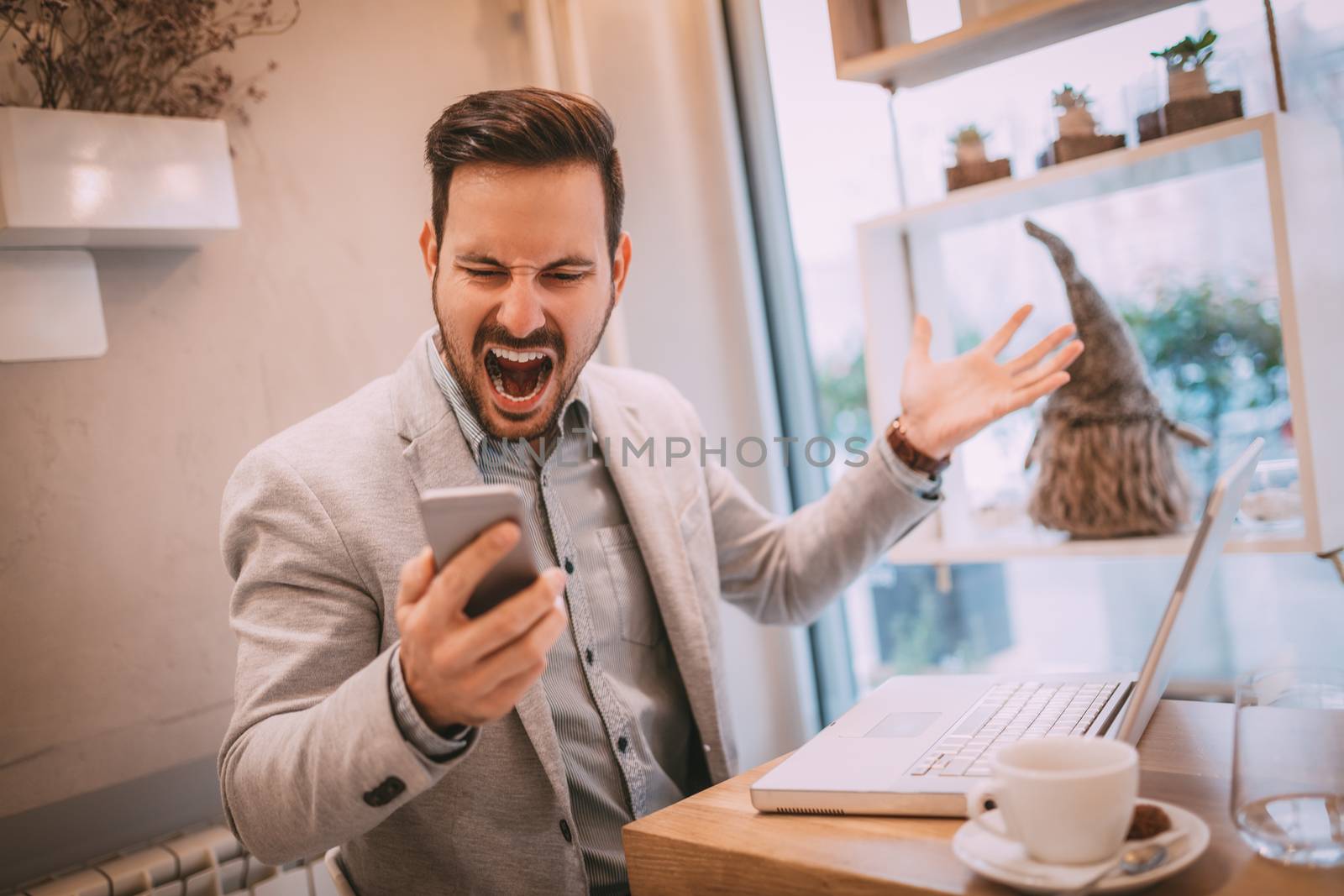 Young stressed businessman screaming on the smart phone and working at the laptop in the cafe.