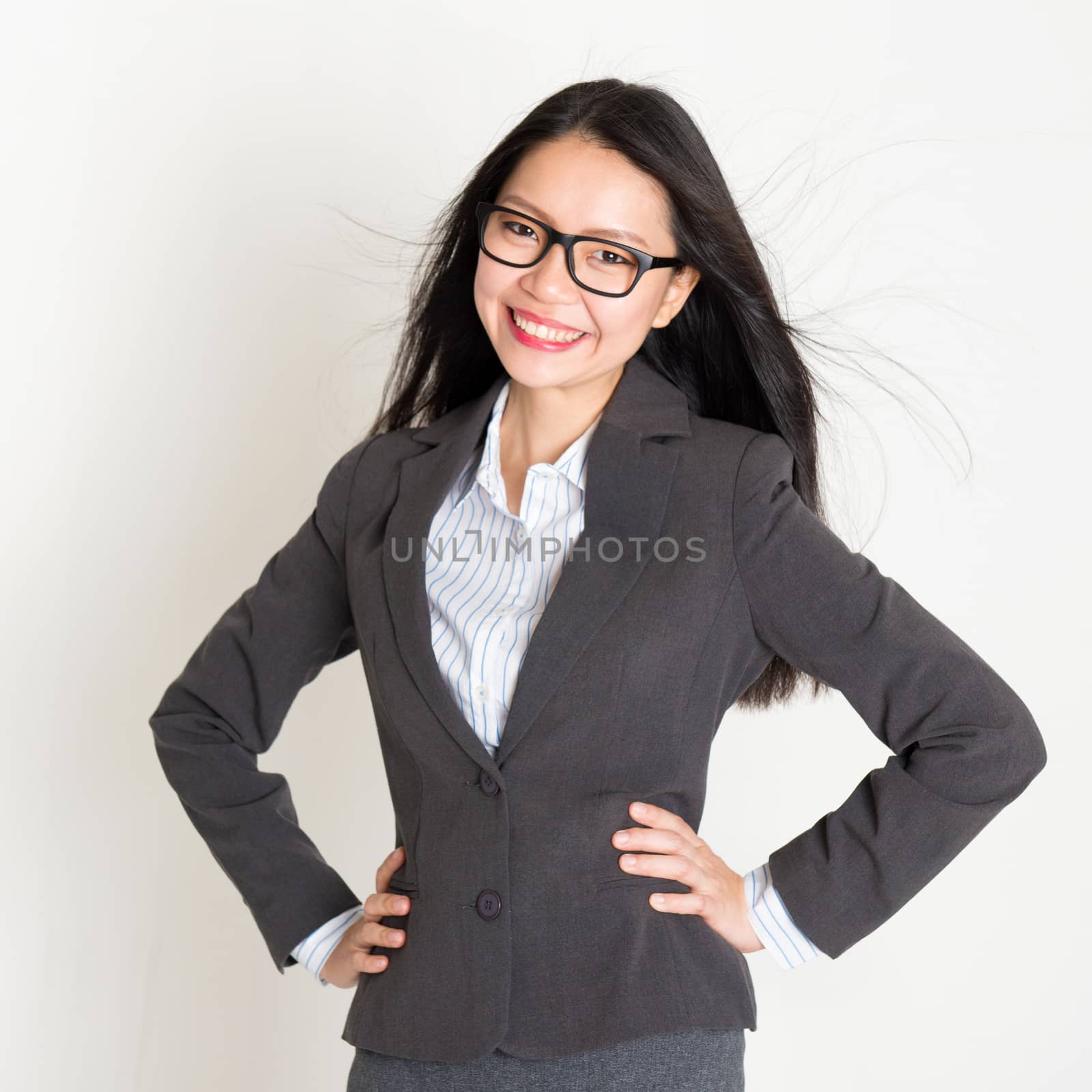 Portrait of young Asian business woman in formalwear smiling and looking at camera, standing on plain background.