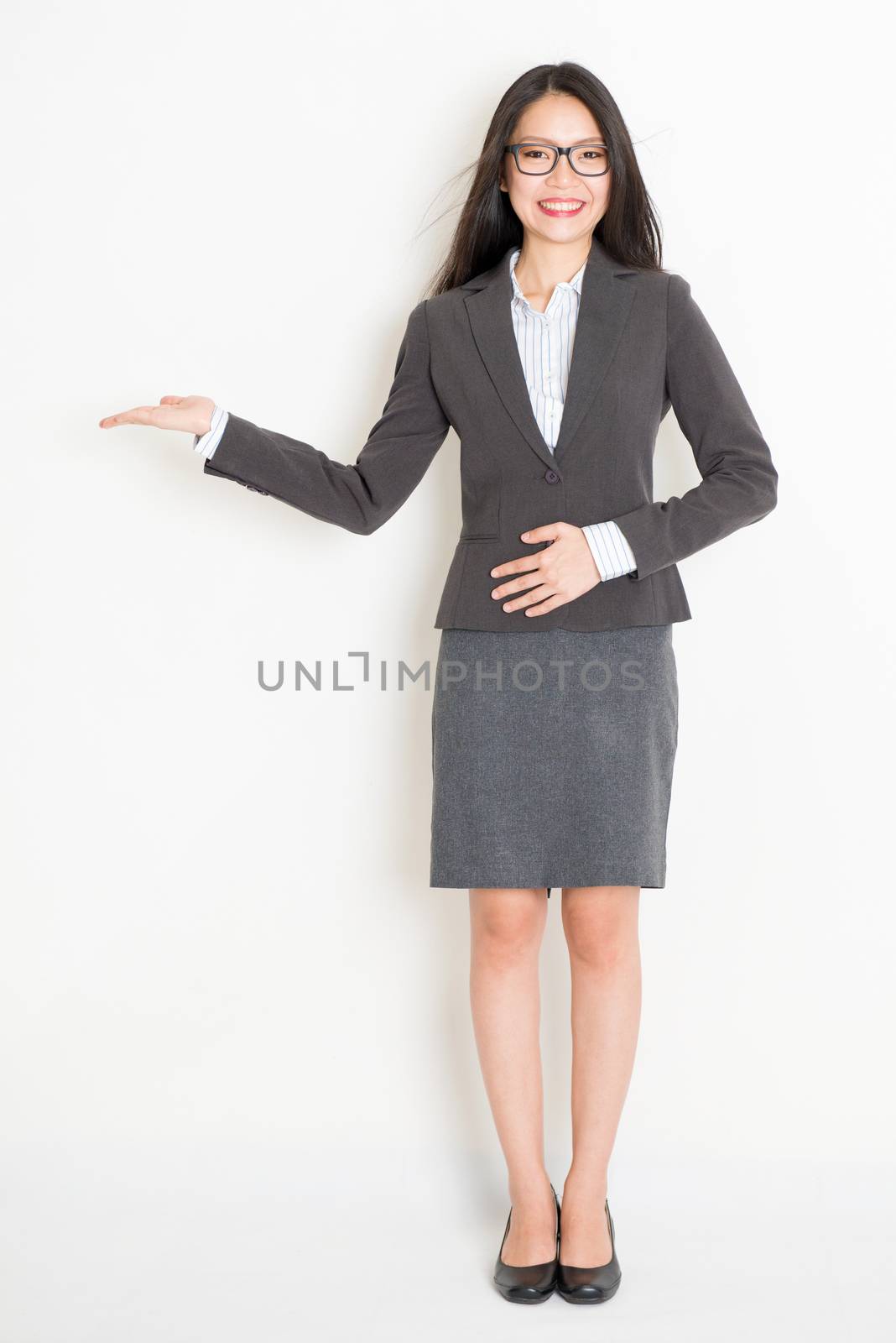 Portrait of Asian businesswoman in formalwear hand showing something and smiling, full body standing on plain background.
