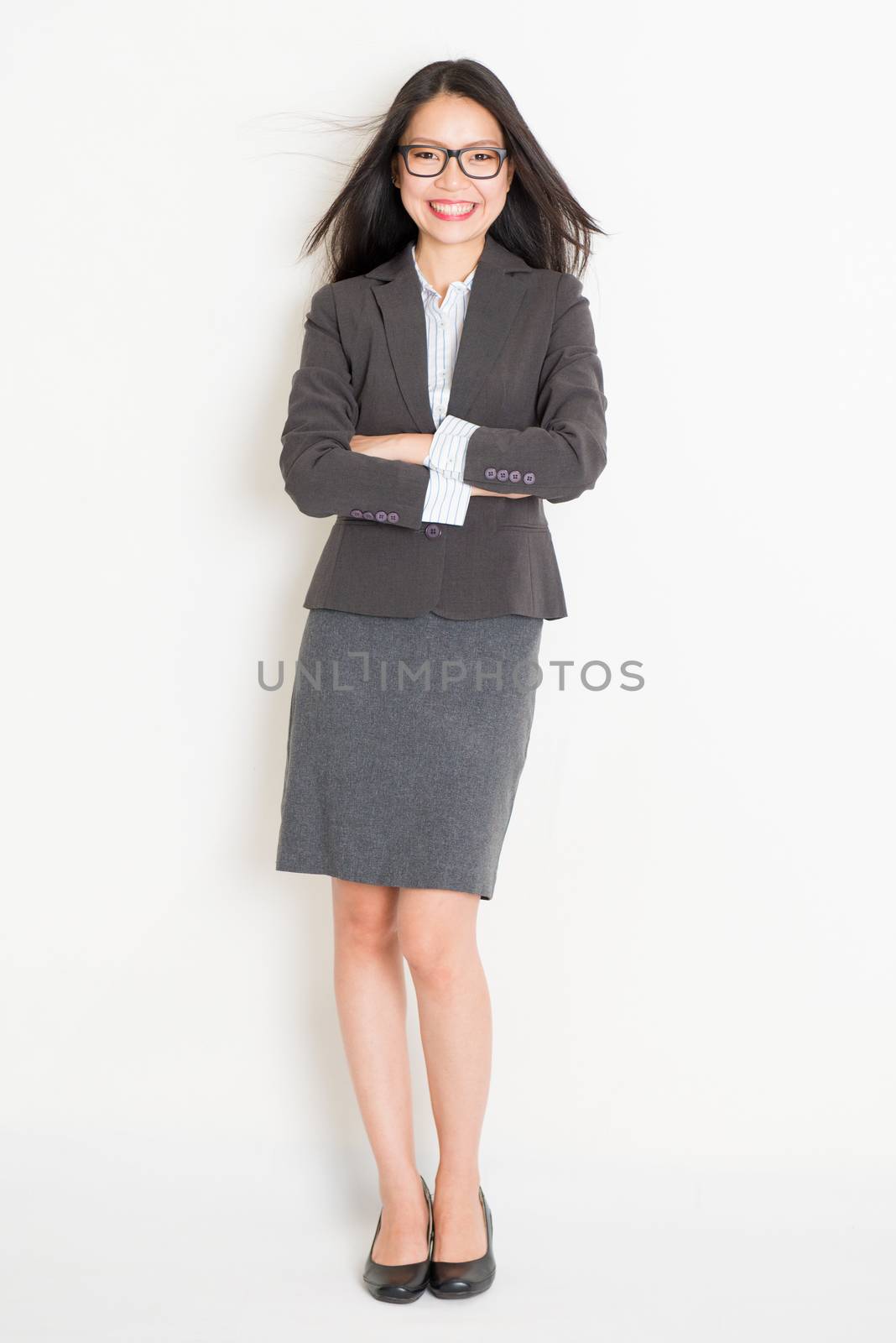 Portrait of Asian female business woman in formalwear smiling, full body standing on plain background.