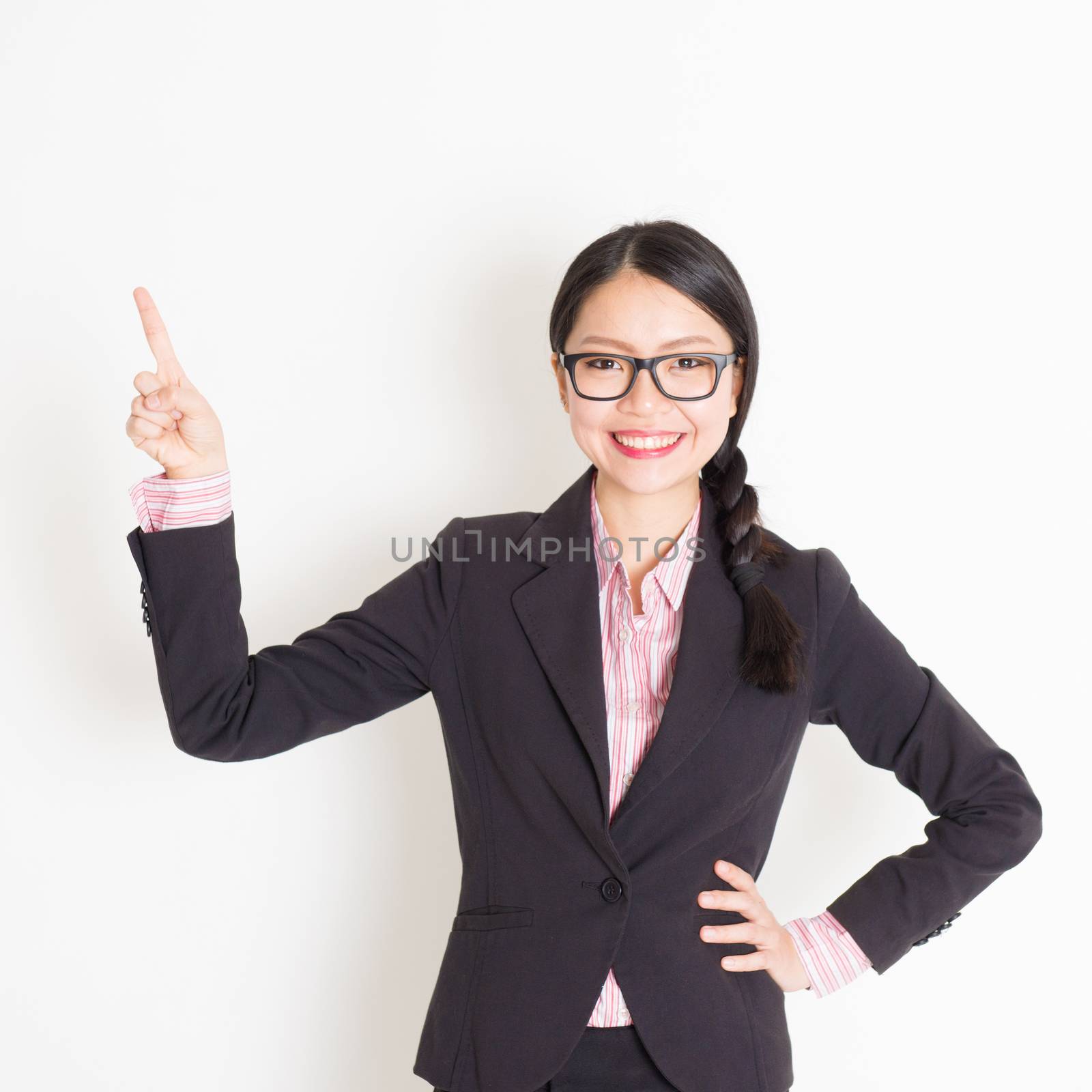 Portrait of Asian businesswoman in formalwear smiling and finger pointing on something, standing on plain background.