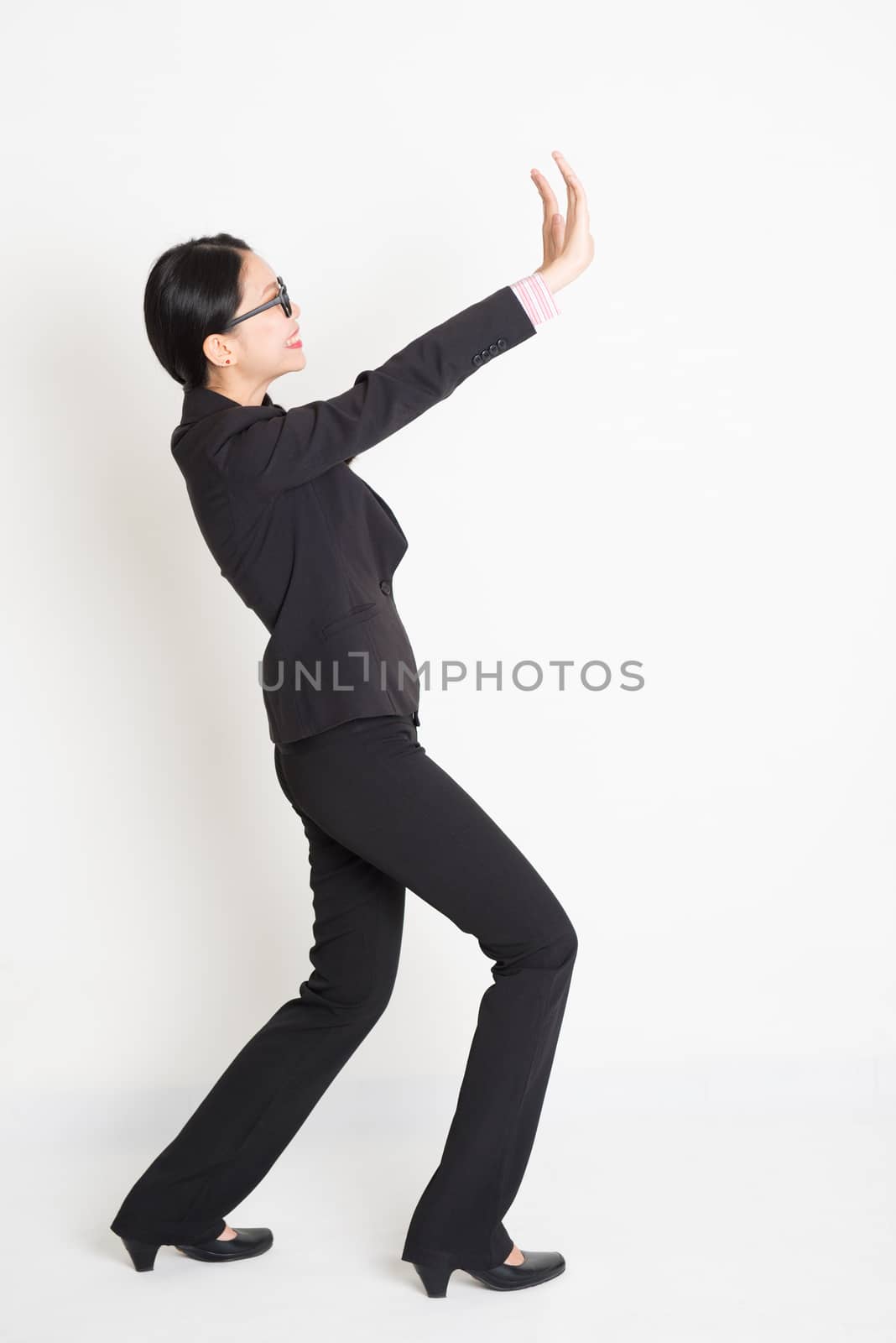 Full body portrait of young Asian businesswoman in formalwear hands pushing on blank space and smiling, standing on plain background.