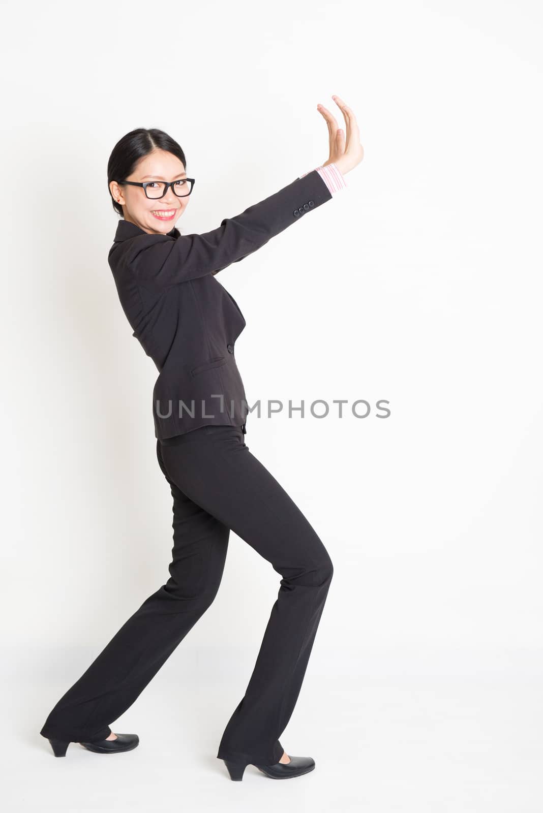 Full body portrait of young Asian businesswoman in formalwear hands pushing on something and smiling, standing on plain background.