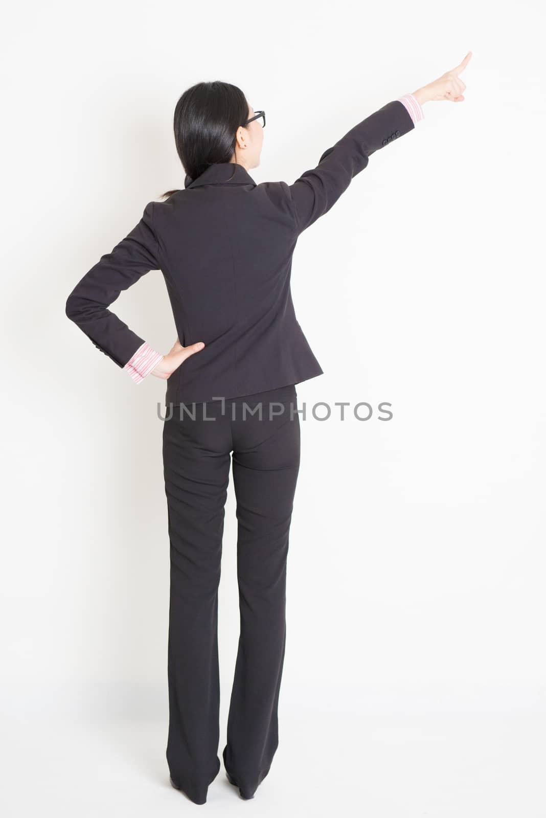 Full body back view of young Asian businesswoman in formalwear finger pointing on something, standing on plain background.