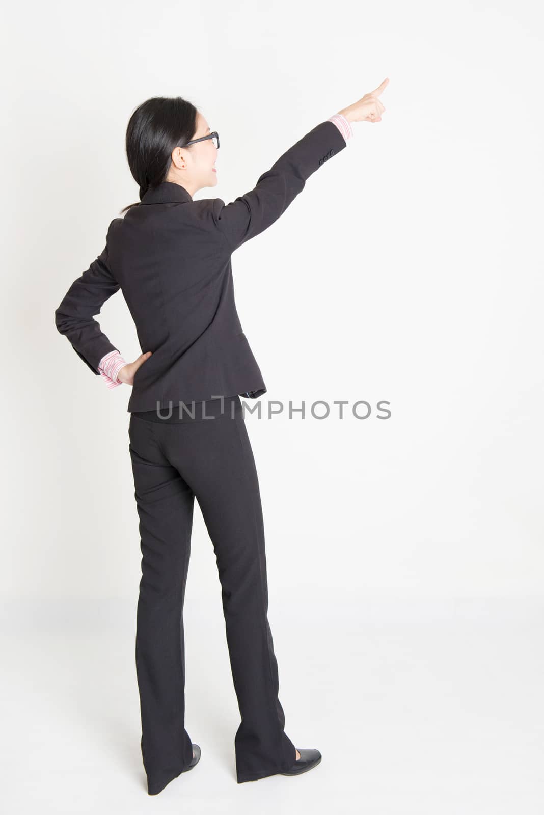 Full body rear view of young Asian businesswoman in formalwear finger pointing on something, standing on plain background.