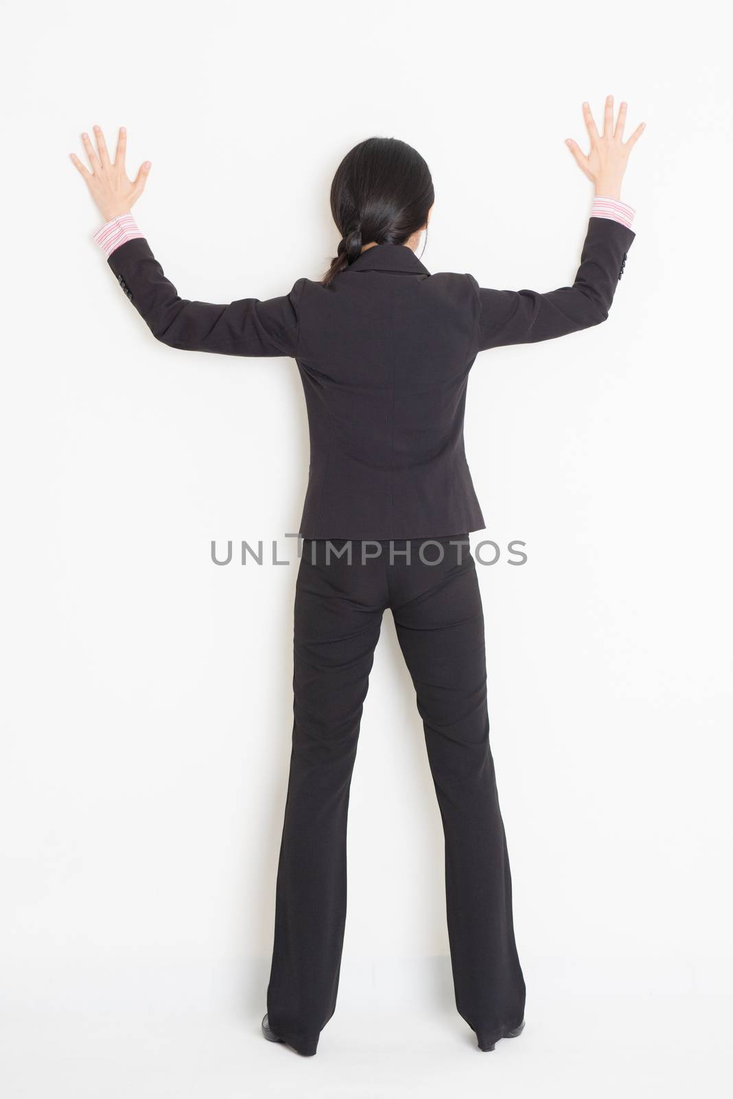 Full body back view of young Asian businesswoman in formalwear banging on wall, standing on plain background.