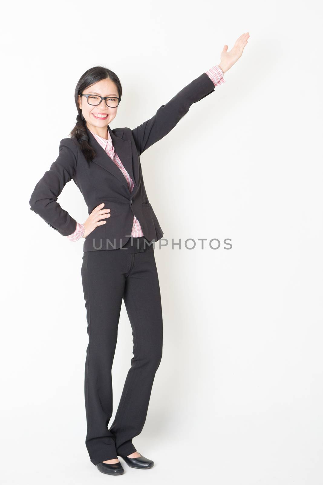 Full length front view of young Asian businesswoman in formalwear hand presenting something, standing on plain background.