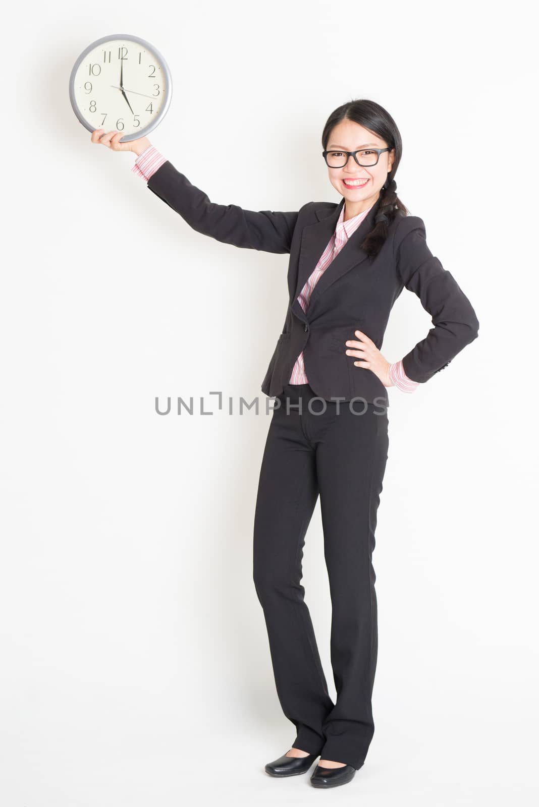 Full length front view of young Asian businesswoman in formalwear hand holding clock at 5pm, standing on plain background.