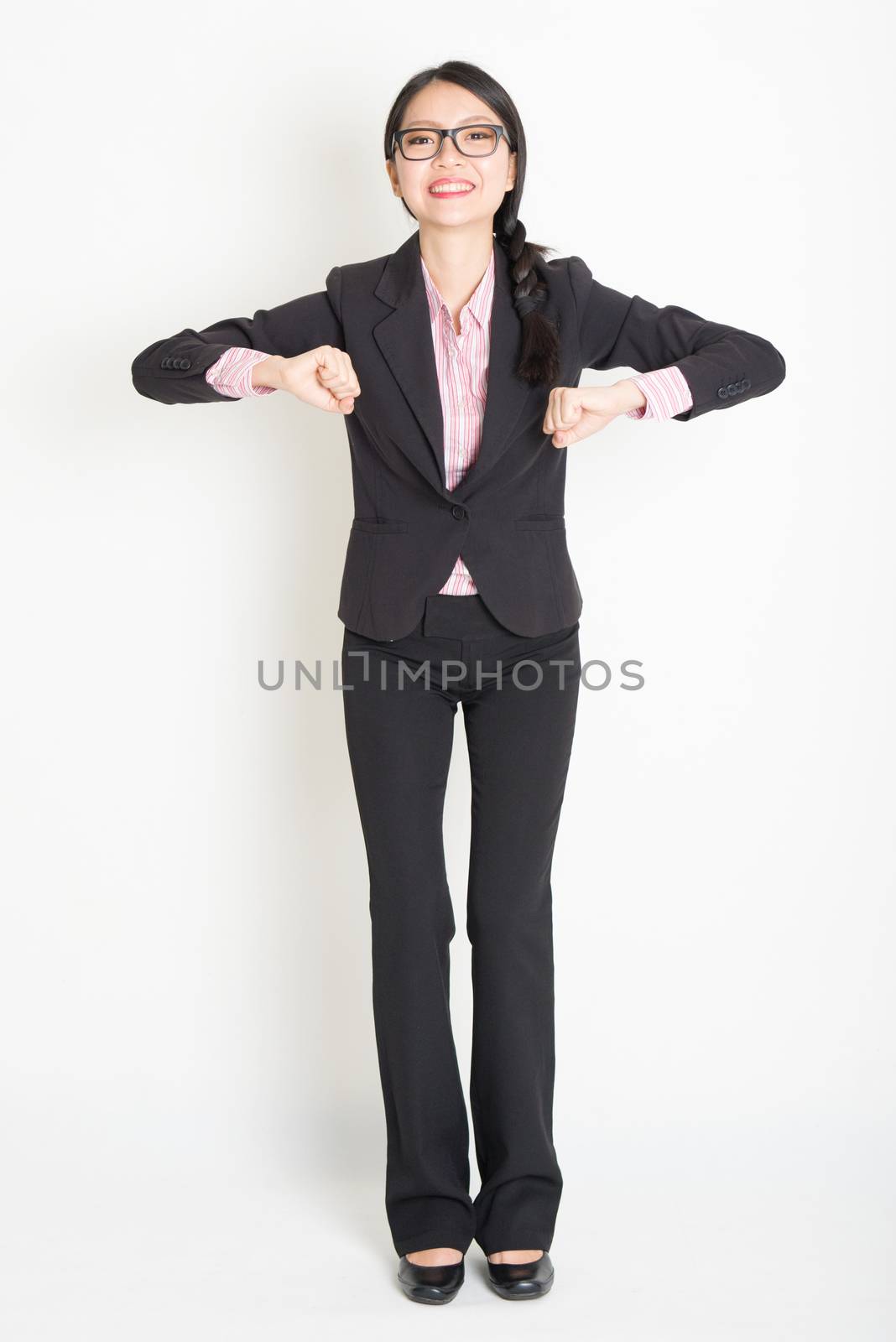 Full length front view of young Asian businesswoman in formalwear ready to jump, standing on plain background.