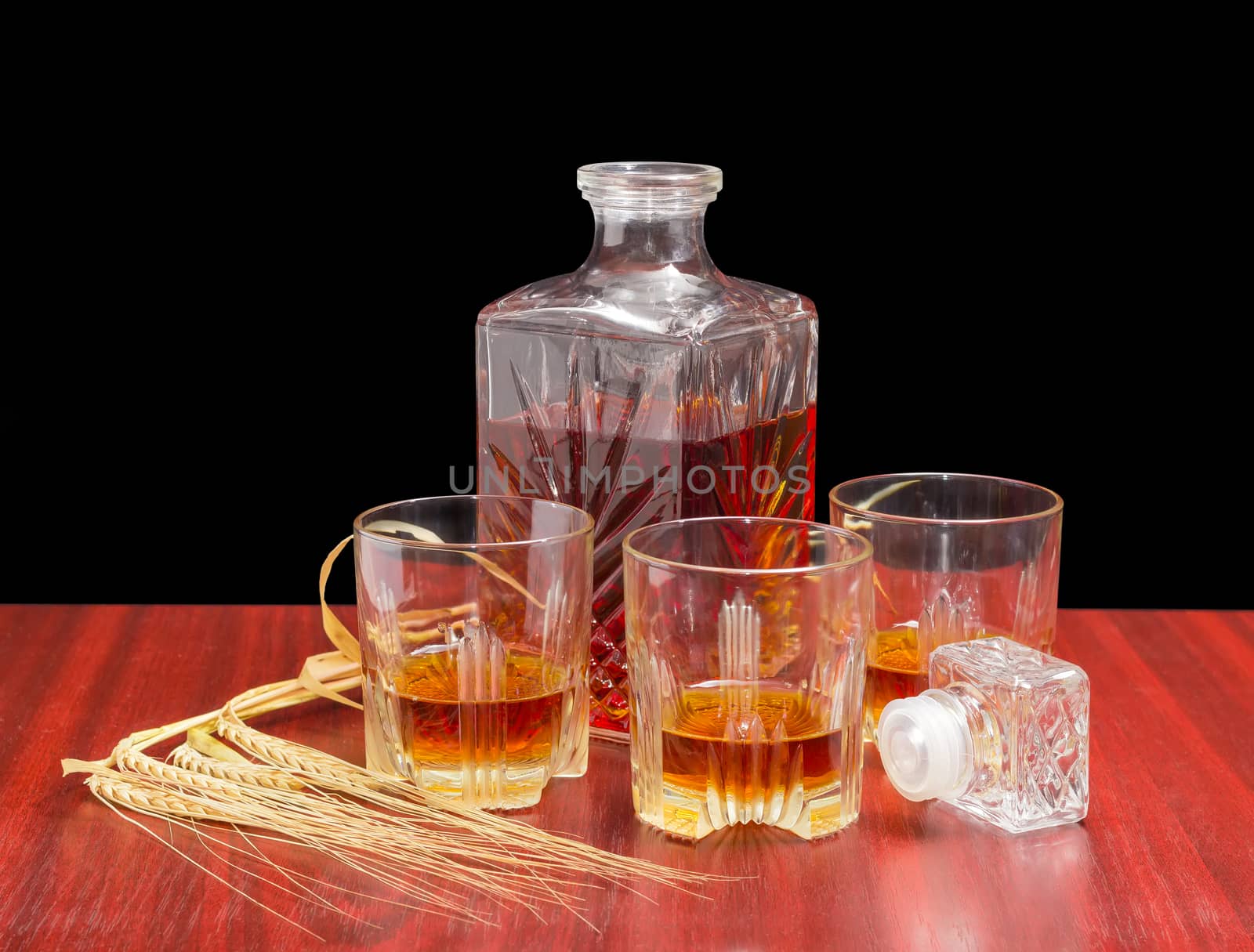 Decanter and three glasses with whiskey, stopper from the decanter and several barley spikes on a wooden table on a dark background
