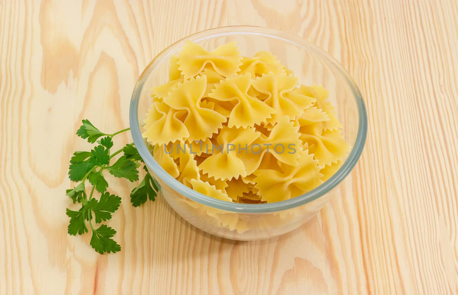 Uncooked bow-tie pasta in glass bowl and sprig of a parsley on a light wooden surface
